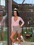 A woman posing in the Be Athletica Ultimate Ballet Wrap Top in Mauve standing in front of a glass door at a padel court.
