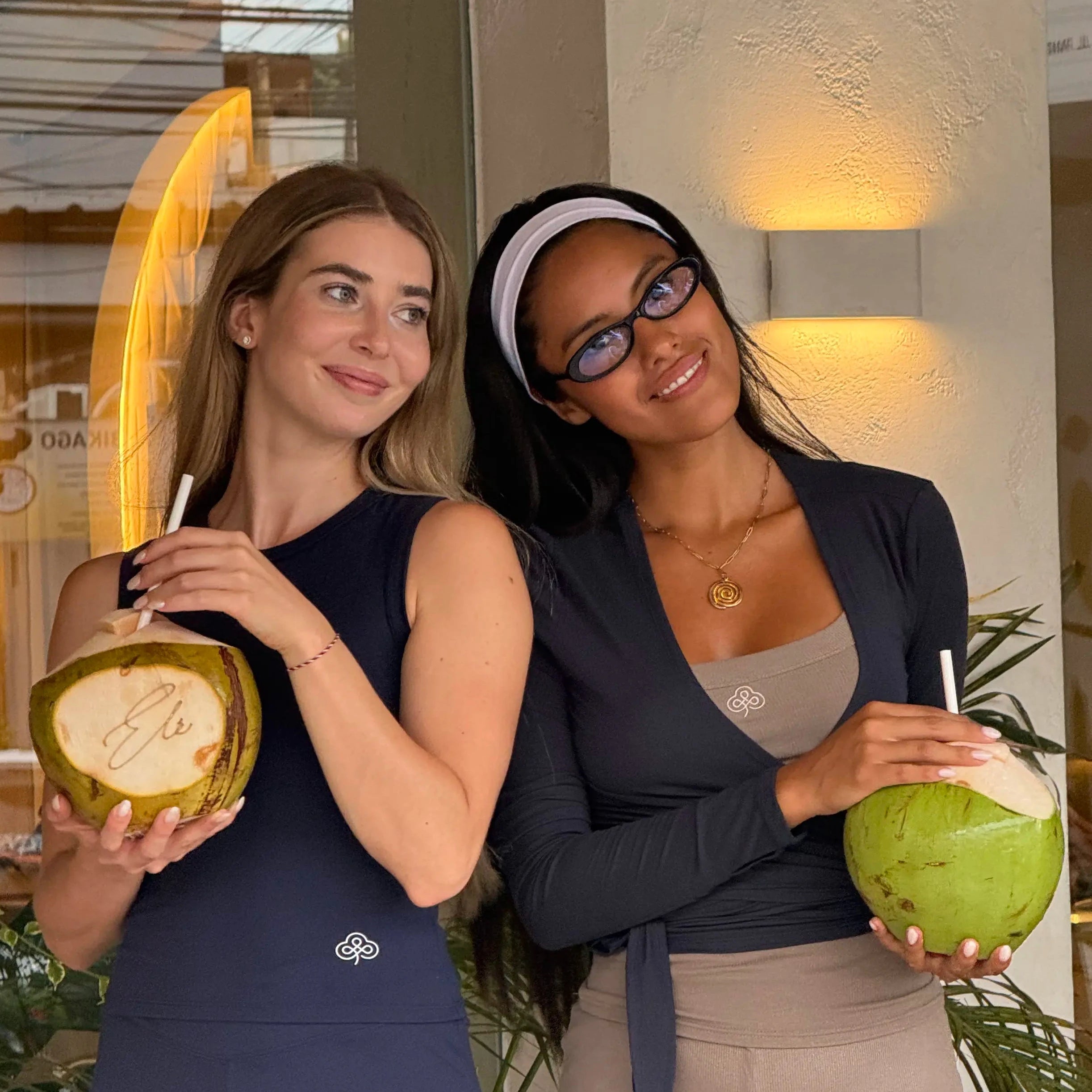Two women posing together in a cafe setting, each holding a coconut with a straw. The woman on the left wears a Be Athletica The Ultimate Ballet Wrap Top in Midnight and matching leggings, while the woman on the right wears a Be 180 Classic Tank in Midnight, beige shorts, and a white headband.