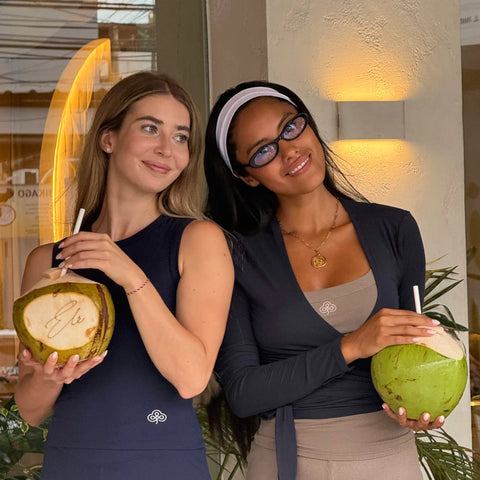 Two women posing together in a cafe setting, each holding a coconut with a straw. The woman on the left wears a Be Athletica The Ultimate Ballet Wrap Top in Midnight and matching leggings, while the woman on the right wears a Be 180 Classic Tank in Midnight, beige shorts, and a white headband.