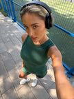 A woman in the Be Athletica Be 180 Classic Tank Top in Forest top and Be 180 Cycling Shorts in Forest wearing black headphones, taking a selfie outdoors on a paved walkway next to a blue fence.