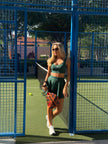 A woman stands on an outdoor padel court wearing a Be Athletica Be 180 Sports Bra and Be 180 Skort in Forest.