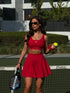 A woman stands on a tennis court wearing a matching Be Athletica Be 180 Sports Bra and Be 180 Skort in Cherry, holding a tennis racket and a tennis ball. The background shows a tennis net, green court, hedges, and a modern building.