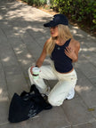 A woman kneels on a paved outdoor walkway wearing a navy blue Be Athletica Be 180 Racer Tank top in Midnight, and Varsity Athletica Wide-Leg Sweatpants in Marshmallow, a navy baseball cap, and glasses. She holds a takeaway coffee cup next to a black tote bag with white text.